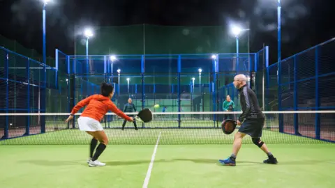 Four people playing the raquet sport padel at night on an artificial grass court. There are two people on one side of the net, one wearing an orange hoodie with white shorts and another with a black jacket and blue shorts. There are two others facing them.