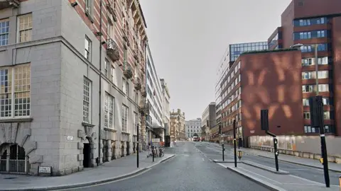 Street view image of James Street. On the left side, a large stone building dominates the foreground. The lower floors are made of light grey masonry blocks with tall, multi-pane windows and arched basement openings. Above, the building transitions into red brick with decorative balconies. On the right side, several modern office buildings line the street. The closest building is a large reddish-brown brick structure.