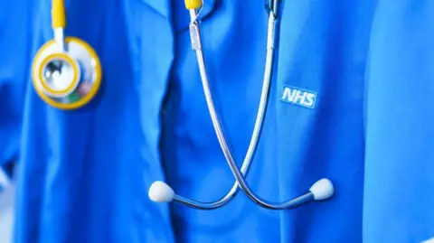 Generic image of blue NHS nurse scrubs. It is a close up of the chest area. There a stethoscope around the neck.