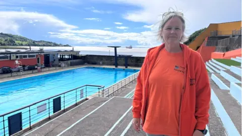 Emma wearing an orange t-shirt and fleece is stood on the pool viewing steps to the left of the picture. The pool and Bristol Channel is visible in the background.