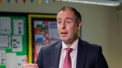 PA Media Paul Givan. He has short grey-ish hair, wearing a navy blazer, white shirt and pink tie. Classroom displays are bring him.