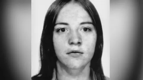 A black and white image of Marian Beattie. She has long hair and is staring straight at the camera in a mugshot-style image.
