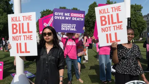 People holding signs saying 'kill the bill not the ill' and 'give me choice over my death' stand protesting outdoors in Parliament Square in early June 2025.