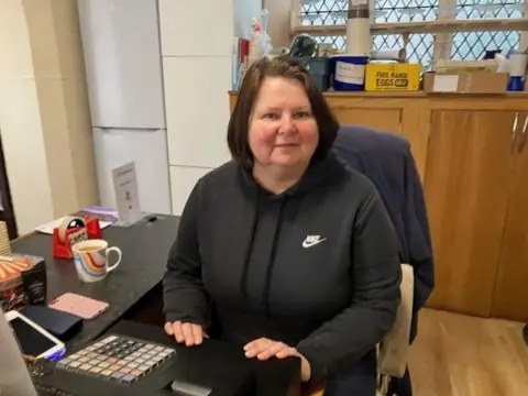 Sheila Donaldson wearing a black hoodie, sat at a till in the cafe of The Strand Centre. Next to her on the side is a cup of coffee. She dark hair, styled in a bob with a gentle smile.