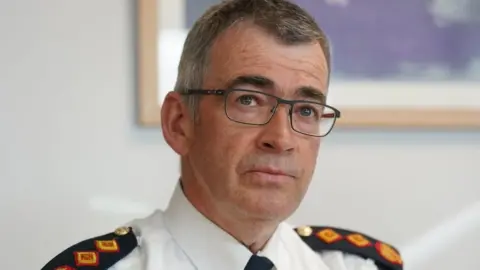 PA Media Drew Harris looks upwards and is wearing glasses. He has short dark and grey hair and is clean shaven. He is wearing a white shirt, black tie and has black, red and yellow shoulder badges with gold buttons.