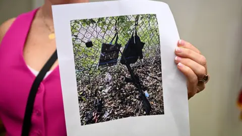 Getty Images A printed photo held by a person wearing a pink top shows the gun and other items, which appear to be bags hanging from a fence, found near where a suspect was discovered during a press conference regarding an apparent assassination attempt on former President Donald. Trump on September 15, 2024, in West Palm Beach