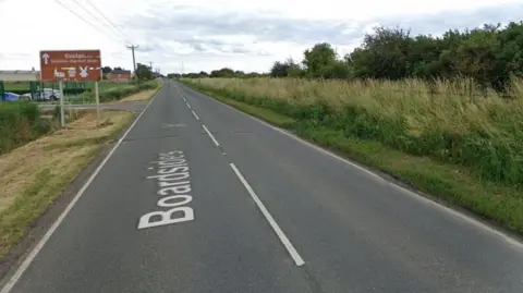 A Google street map image of a road. There is a sign up ahead on the left that reads 'Boston, historic market town' with an arrow next to it pointing ahead. The road is lined either side by grass verges and trees.