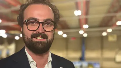 Phil Shepka/BBC Ross Martin stares towards the camera. He wears a white shirt and black jacket. He has a beard and black rimmed glasses. 
