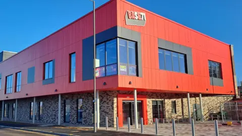 BASE71 An exterior shot of the two-story building, which is cladded in bright red panels and has large windows.