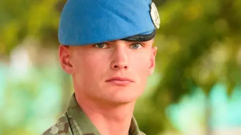 Supplied Liam Potts, a young man wearing a camoflauged tunic and a sky-blue beret with a shining badge on it. He is clean-shaven and looks stoically towards the camera with blue eyes.