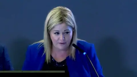 Pauline Hart speaks into a microphone. She has a on a blue blazer and black top. She has blonde hair and brown eyes.