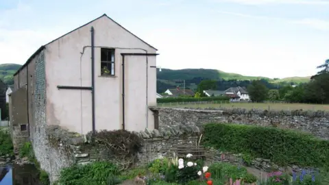 Adam Baker The gable end of Yew Tree Hall. It has a small window on the second floor. The building has a river to the left side. A field is on the other side of a road, and hills can be seen in the distance.
