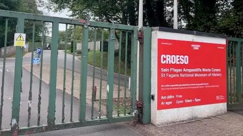  a closed green metal gate, with a red sign to the right with Croeso (Welcome) written in white capital letters and the museum's name and opening times underneath.
