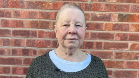 Linda Haigh pictured in front of a brick wall at her home. She has short hair and is wearing earrings and looking directly into camera