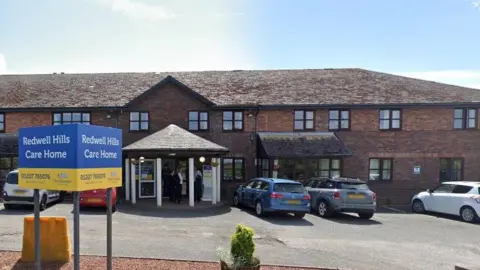 The entrance of Redwell Hills Care Home. It's a long, two-storey, brown brick building. There are five cars parked outside and a sign reading Redwell Hills Care Home.