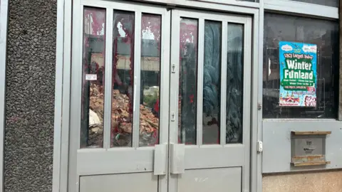 Metal doors with glass windows in them. Through the glass piles of bricks and building bags can be seen. On the wall next to the door is the old night safe hatch.