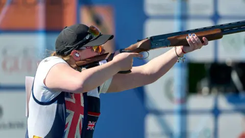 ISSF Bethany Norton holding and aiming her gun. The picture is taken side on. She has a black cap on her head.