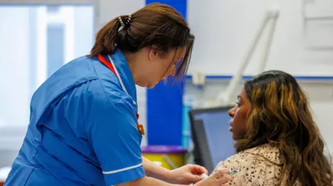 Liam McBurney/PA Wire A nurse, dressed in uniform, is giving a vaccination to a patient. The nurse is standing to the left of the patient who is sitting down. She is wiping the patient's arm with cotton wool. They are inside a hospital.