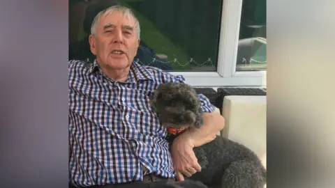 Reg Bown Reg wearing a shirt and holding a small black, fluffy dog.