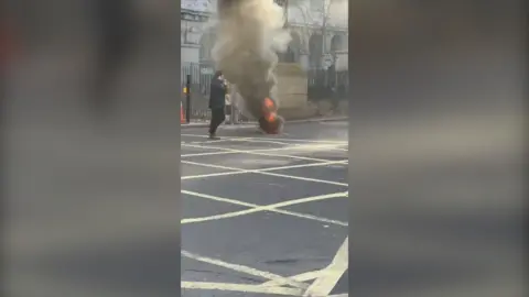 Thick smoke billows from a vent in the middle of a London road. A person walks near to it while filming on their phone.