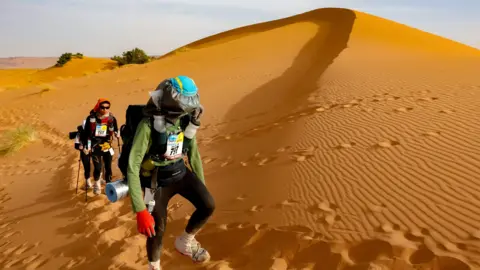 Getty Images Three people in running gear climb a sand dune in the Sahara while taking part in the Marathon de Sables, surrounded by sand covered in footprints left by other runners.