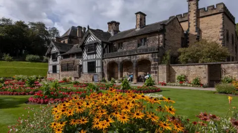 Getty Images A large historic building with black and white timbered sections as well as blackened stonework sit in a park with flowers and lawns