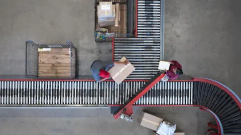 Getty Images An overhead, bird's-eye view shows two logistics workers operating a warehouse conveyor belt system on a concrete floor. One worker wearing a grey shirt and a red cap leans over to lift a cardboard box from the main horizontal roller conveyor. Across from them, another worker in a maroon long-sleeve shirt stands holding a yellow clipboard. The automated conveyor system consists of metal rollers framed in red, with a curved section leading off to the bottom right. Nearby, several large wire storage cages contain packaged goods, and loose cardboard boxes rest on the floor.