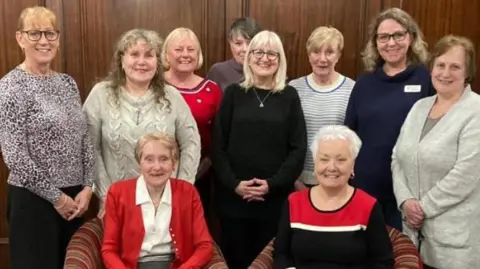 A group of ten women in a posed photo. Two are sitting down in armchairs and the others stand around them in front of wooden panelling.