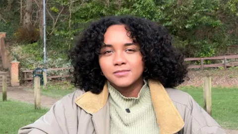 Chris McHugh/BBC Julia Anyanwu poses for the camera in a Maidenhead park. She has long curly black hair, and is wearing a beige jacket with a green roll neck jumper underneath. A wooden fence and trees can be seen in the background.