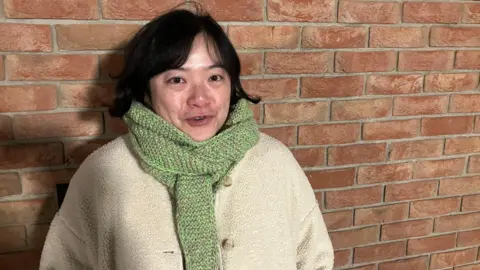 Amy Holmes/BBC A woman wearing a light, textured coat and a green knitted scarf stands in front of a brick wall.