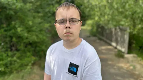 Alfie Ford wearing a white t-shirt. He is wearing glasses. We can see there is greenery behind him.