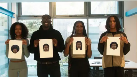 Four young people - two male and two female -  stand in a line holding up pieces of paper with their final computerised emoji designs - featuring Afro, cornrow, braids and locs hairstyles