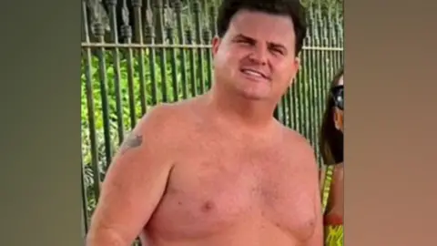 Thomas Casey stands in front of metal railings with greenery behind him. He is topless, and has a small tattoo on his right upper arm. He has dark hair and is smiling at the camera.