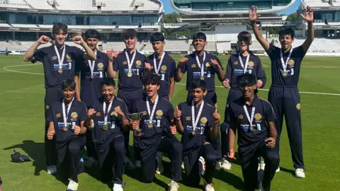 Twelve members of an U15 cricket team, dressed in navy, on the ground at Lord's. They are all wearing medals and have either thumbs up or fists raised. One member is carrying a cup.