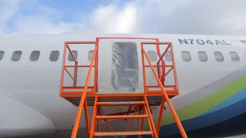 NTSB The exterior of a Boeing jet is show with plastic sheeting covering an open gap where the door was. A ladder leads up to it. The plane is white with a multicoloured stripe. 