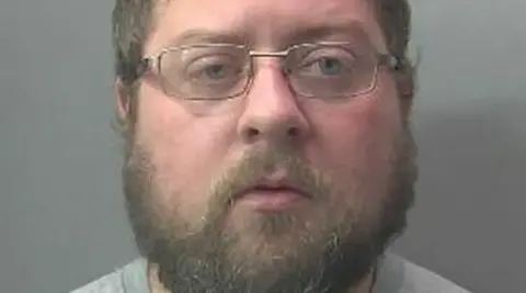 Cambridgeshire Police A mugshot of a man who has facial hair and glasses. He is looking into the camera. 