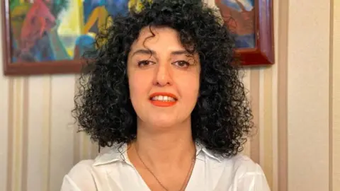 A woman with curly black hair dressed in a white collared shirt and red lipstick, smiles to the camera. 