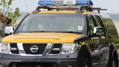 BBC/Dale Baxter Two people sit in the front of a Nissan 4 by 4 vehicle with "HM Coastguard" written on the bonnet and blue lights on the roof, with the word "rescue".