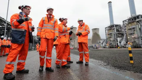 PA Media Four men wearing orange hi-vis clothing stand outside inside a petrochemical plant