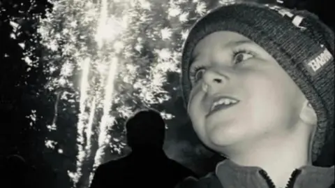 A black and white photograph of Archie York. He is wearing a woolly hat and looking at some fireworks in the distance.