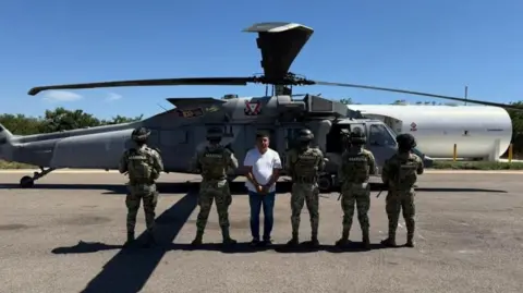 Members of Mexican Special Forces escort Audias Flores, known as “El Jardinero after his capture. Five officers are standing with their backs to the camera, to conceal their identities. Flores is facing the camera. He is wearing a white T-shirt and blue jeans. His hands are cuffed in front of him. Behind him is a helicopter. 
