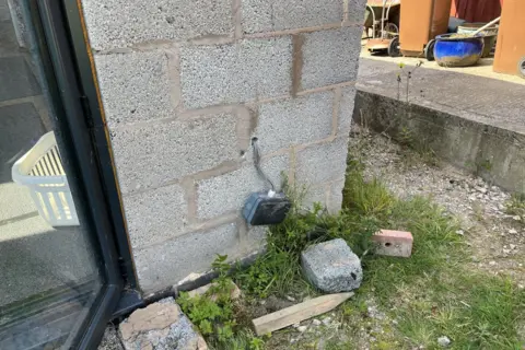 Close up of a corner of a building that has exposed grey breeze block and building rubble on the ground. There is an outside electrical socket dangling off the wall.