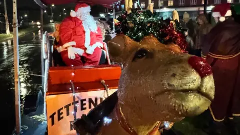 Tettenhall Rotary Club Santa is wearing a red suit and sitting on a sleigh. There is a plastic reindeer at the front of the sleigh with tinsel on its head.