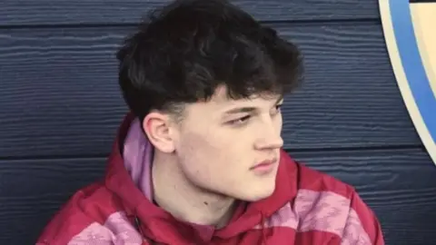A close up image of Alex in the dug-out with black wooden panels behind him. He is wearing a red and pink hoodie. He has dark hair and is looking away from the camera with an intense expression.