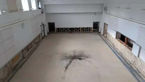 A hall with a large black stain in the middle, white walls and windows high on the left hand side