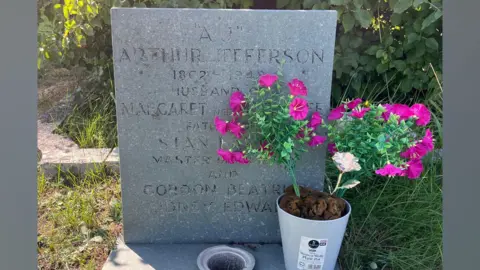 Karl Bird/BBC Headstone for Arthur Jefferson - a grey rectangular stone block with slightly faded inscription. The epitaph reads: Arthur Jefferson, husband of Margaret and father of Stan Laurel, master of comedy. There is a vase of pink flowers to the right.