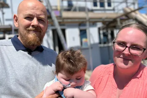 Family photo Amanda and Nick are holding Jack in front of their house which is covered in scaffolding. Amanda has a pink long-sleeved top, wears glasses and has her fair hair tied back in a pony tail. Nick has a beard and is wearing a grey jumper and jeans. Jack is in a blue baby grow and has a feeding tube going up his nose.