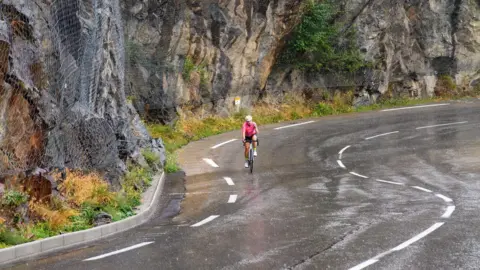 Joolz Dymond A woman cycling far away in the middle of a winding road with a rock wall and wet road