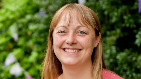 Warwickshire County Council A woman with long brown hair wearing a pink dress and smiling. She is stood in a garden