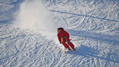 A skier dressed in red skiing gear going down a snowy slope. He is wearing a hat and ski goggles and is carrying poles. There is a cloud of snow behind him. 